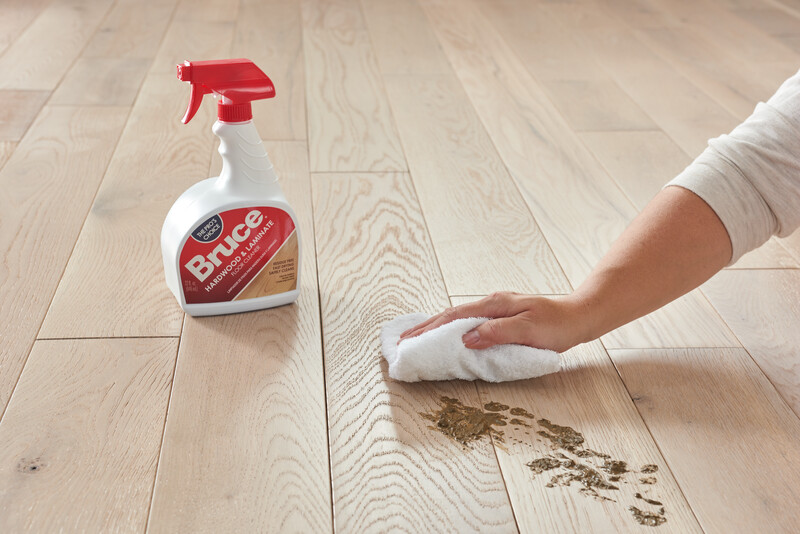 Woman wiping mud from a hardwood floor with a bottle of Bruce Hardwood Floor Care