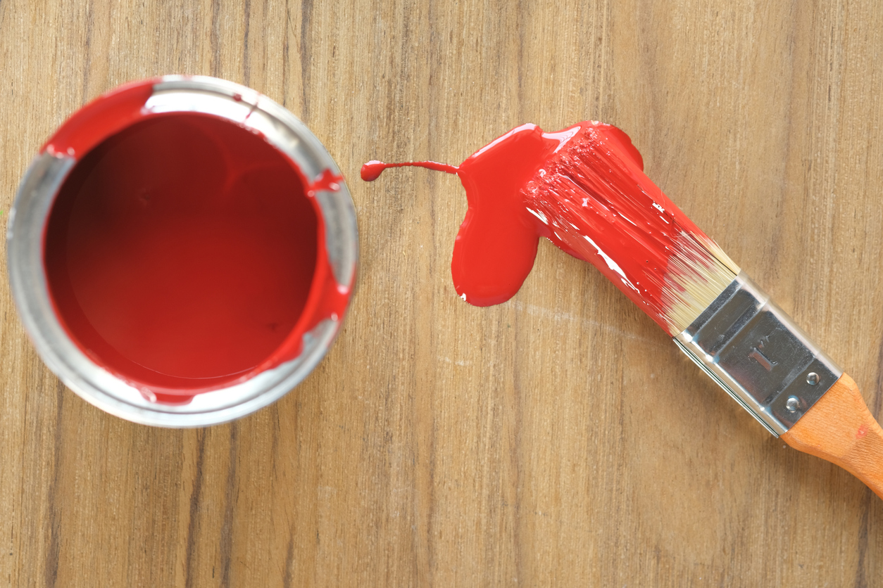 How to remove paint from a hardwood floor. Image shows a red paint can with a red paint brush and paint coming off the brush onto a wood floor.